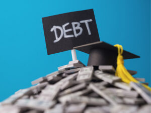 Sign reading "DEBT" on top of a pile of money with a graduation cap.
