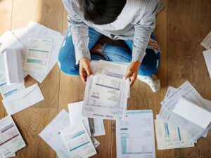 Woman sitting on the floor holding a utility bill with invoices and bills surrounding her.