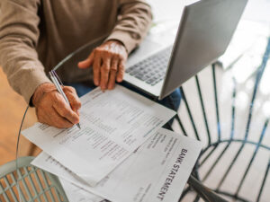 Man sitting at a table reviewing a paper bank statement with a laptop open.